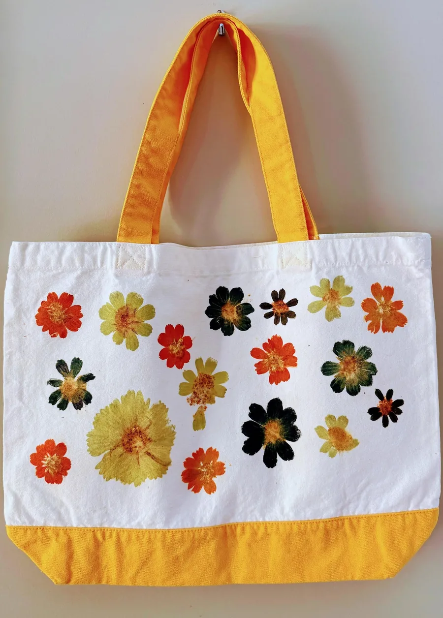 Finished pound-dyed tote bag hanging on a wall hook: white canvas with marigold-yellow handles and base, covered in pressed orange, chartreuse, and deep green cosmos prints