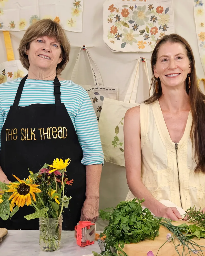 Theresa Brown and Kristi Thompson smiling behind a table of fresh flowers and herbs, with their printed totes hanging behind them