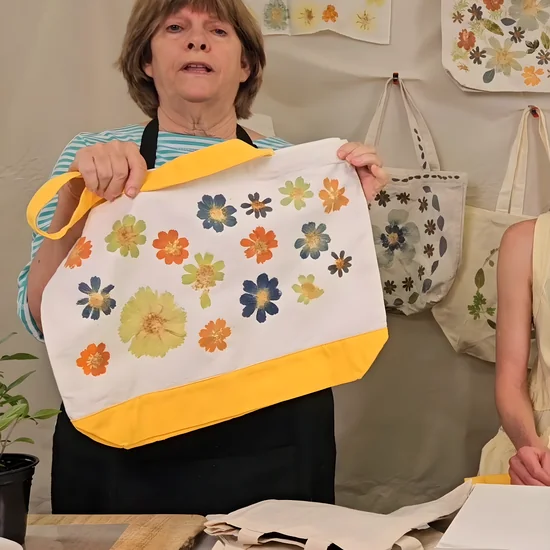Module 1: Theresa holding up a finished tote printed with blue, orange, and chartreuse cosmos in front of the wall of printed bags